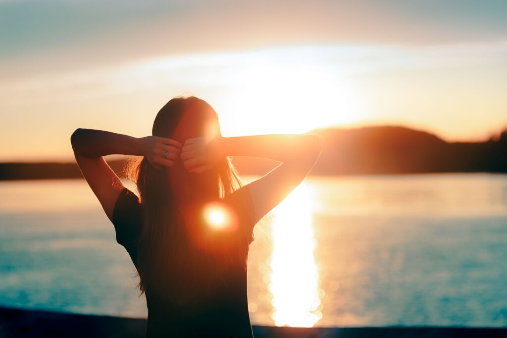 Woman facing sunset over the water with hands behind head