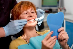 Woman smiling into handheld mirror in dentist's chair admiring smile.